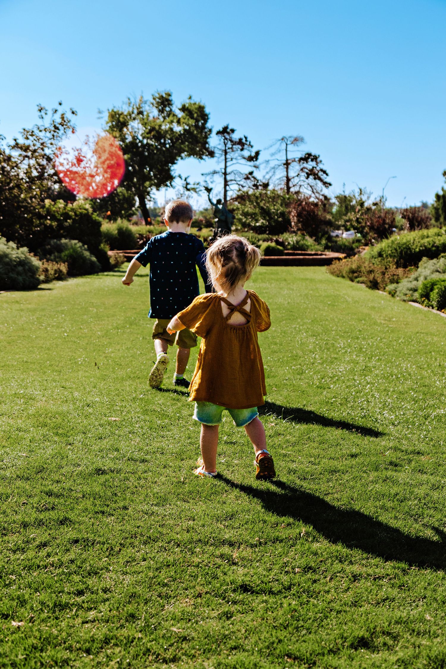 Two young children running on lush green grass in a sunny park, one holding a red balloon.