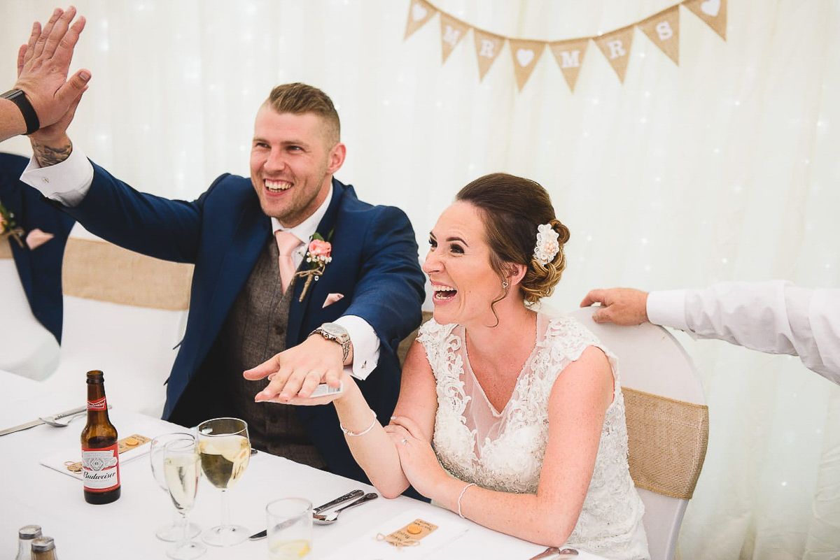 Bride and groom laughing joyfully at wedding reception table, groom in navy suit, bride in lace dress.