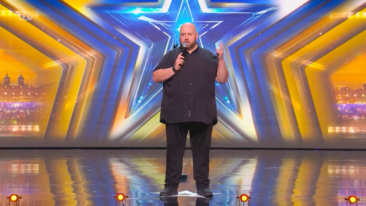 Comedian performing stand-up on Britain's Got Talent stage with golden star backdrop.