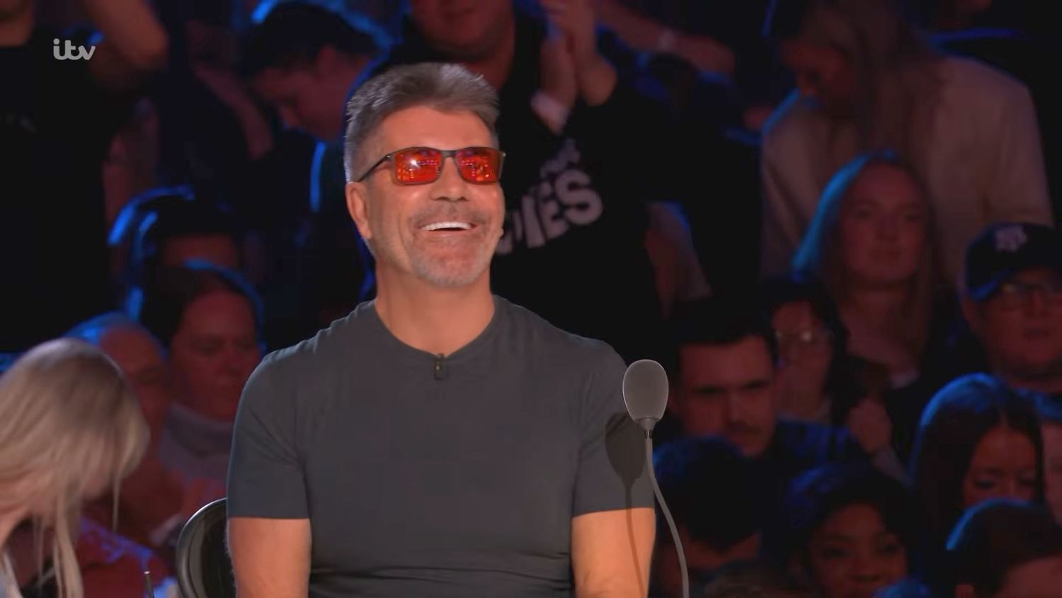 Simon Cowell smiling and wearing orange tinted glasses while seated as a judge on Britain's Got Talent.
