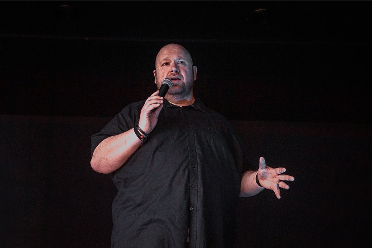 Man in black outfit performing stand-up comedy on stage, speaking into a microphone with expressive hand gesture.