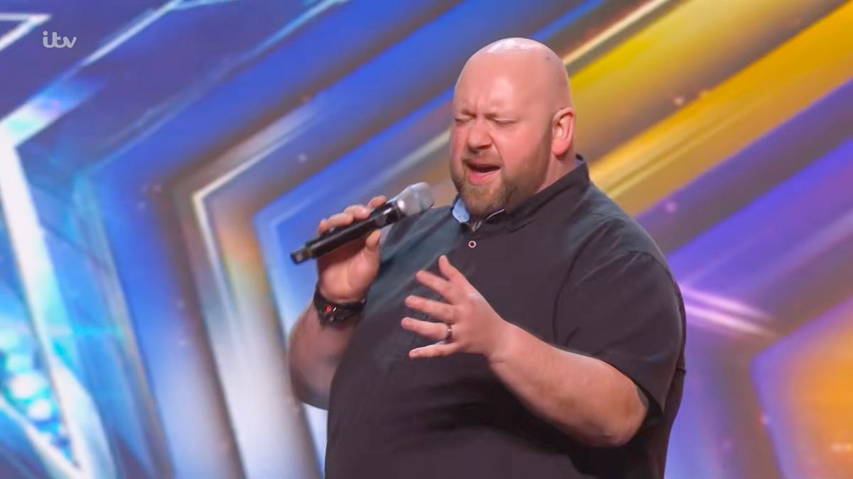 Heavyset man performing on a talent show stage, singing emotionally into a handheld microphone.