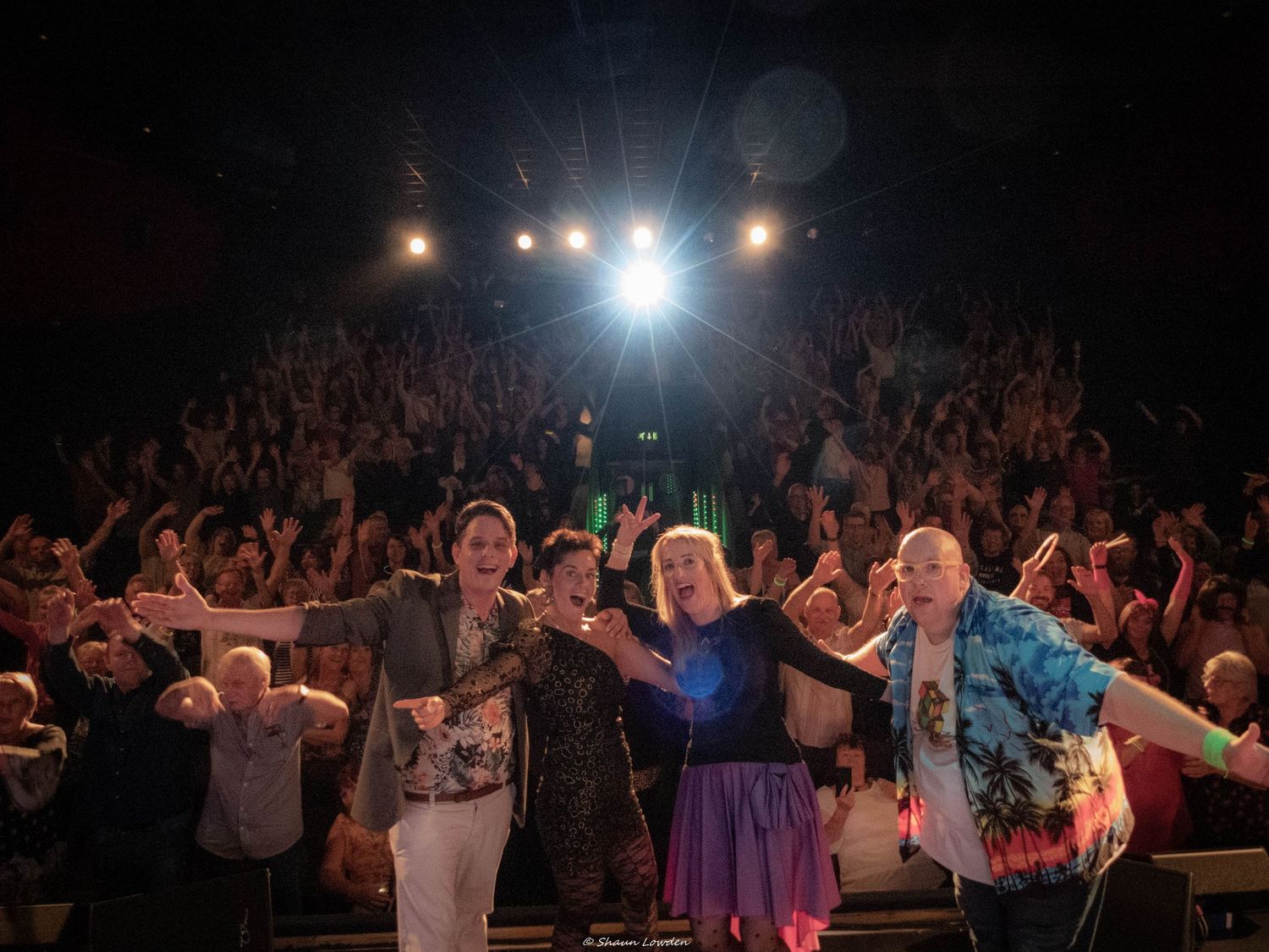 Large crowd of concert attendees with raised arms enjoying a live performance under stage lighting.