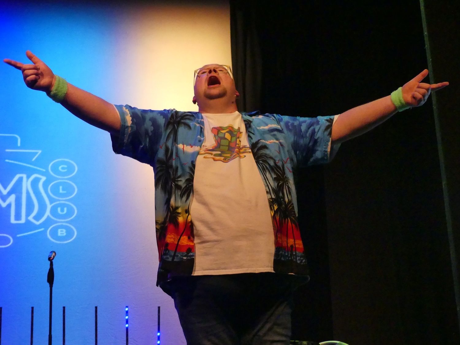 Performer on stage wearing a tropical print shirt with arms outstretched under blue lighting.