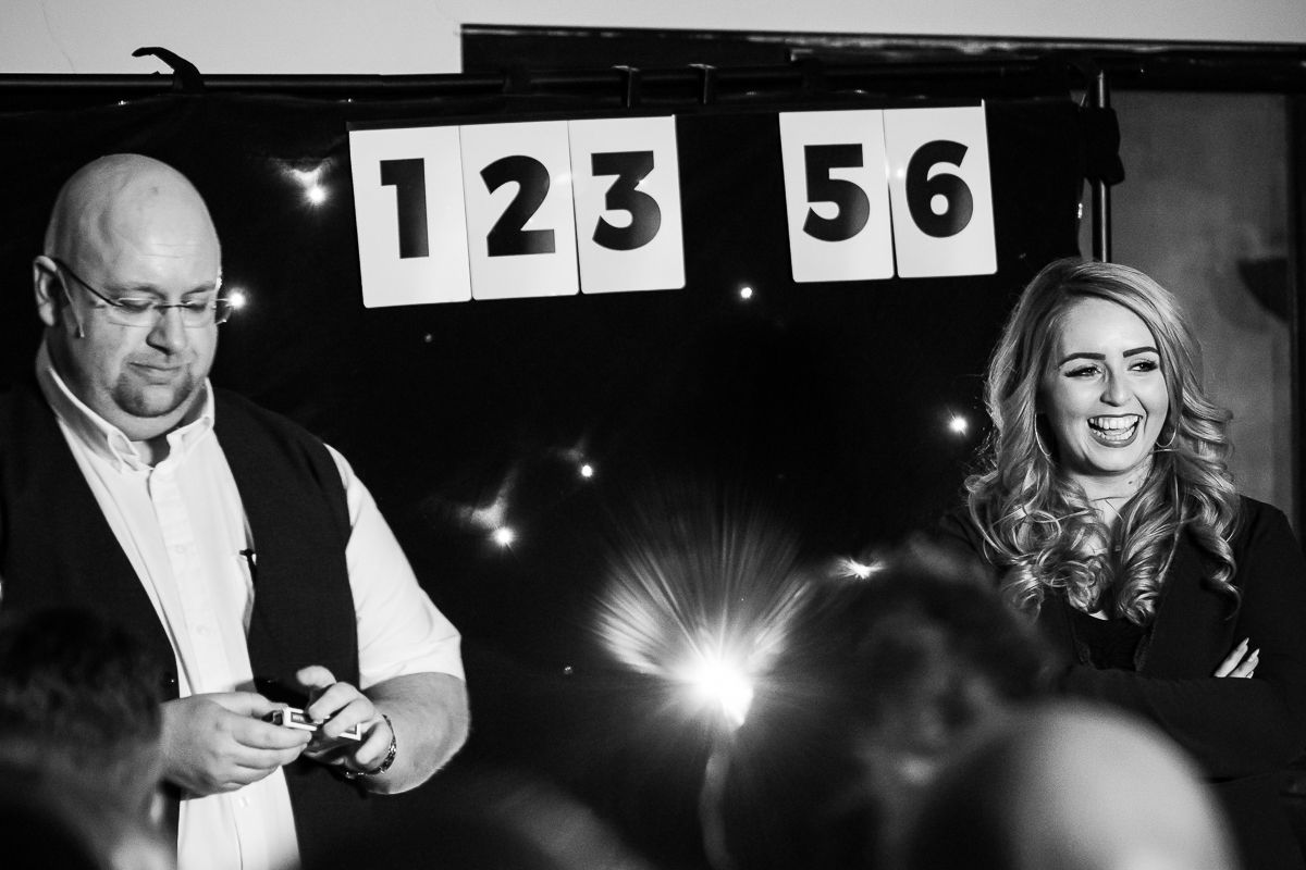 A bald man and blonde woman perform magic on stage with numbered signs in a black and white photo.