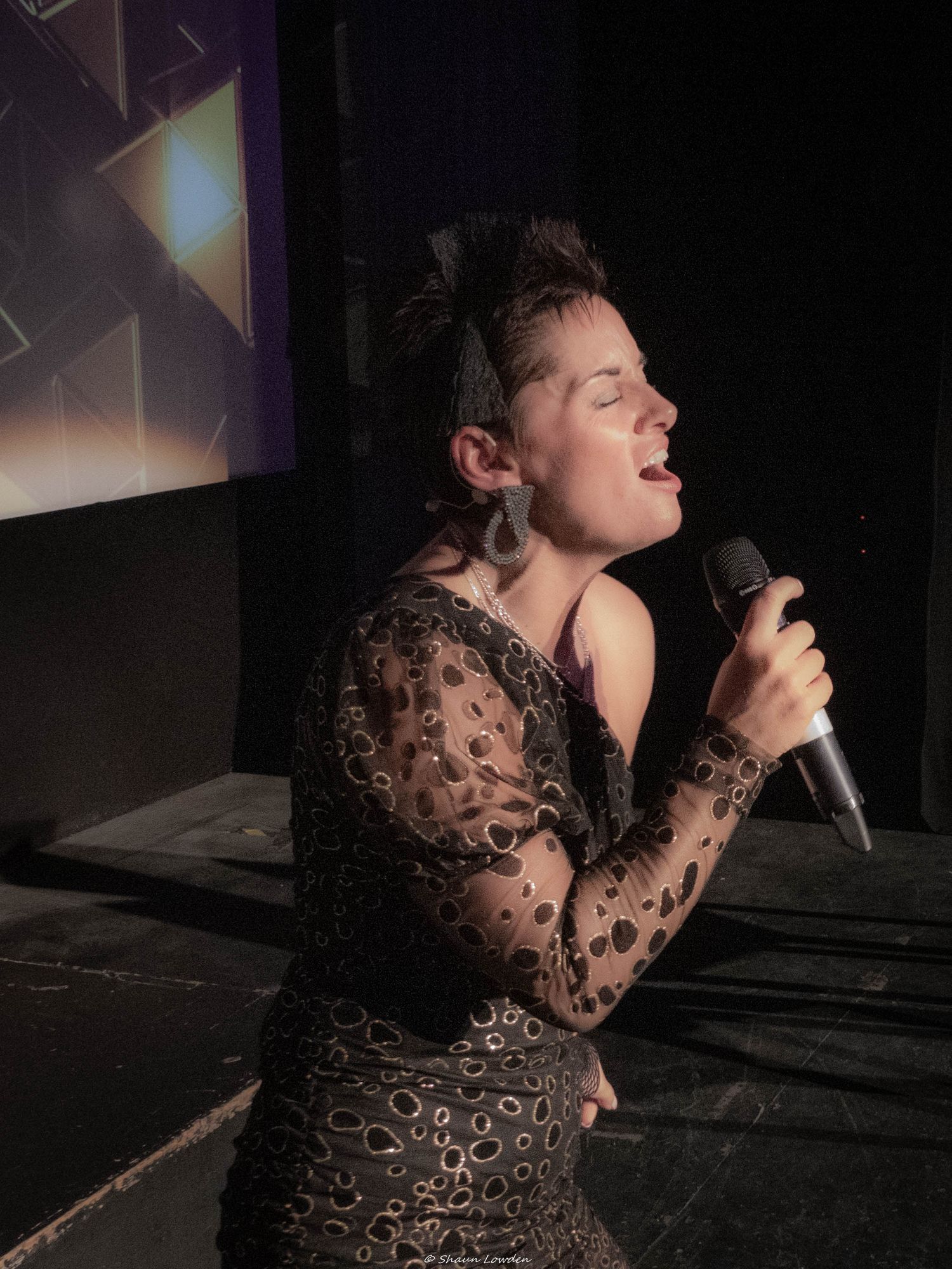 Singer in black lace dress performing passionately on dimly lit stage.