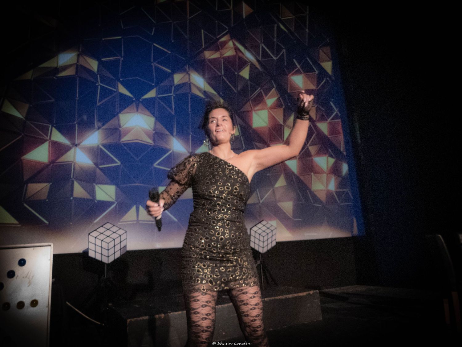 Performer in sparkly dress dancing energetically on stage with geometric patterns in background.
