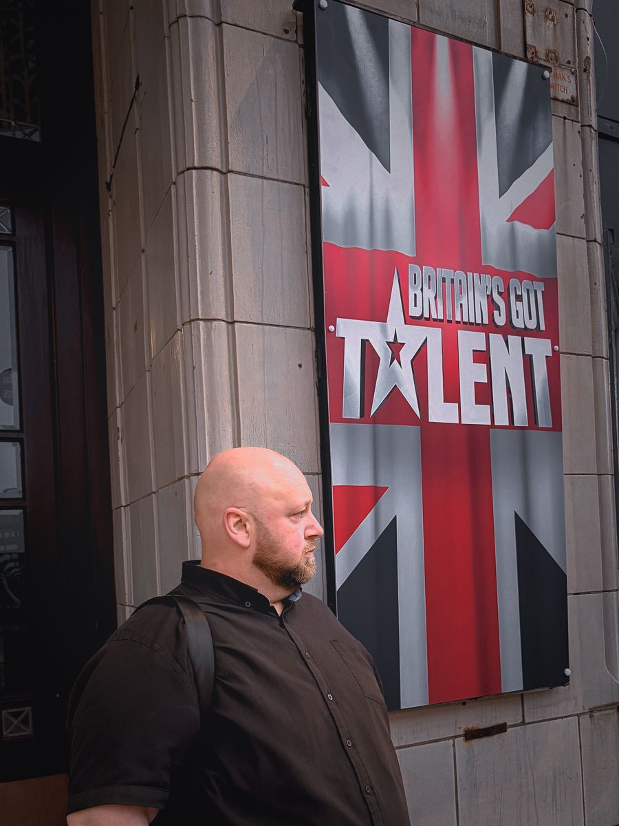 Bald man in black shirt standing beside a Britain's Got Talent promotional banner with Union Jack design.