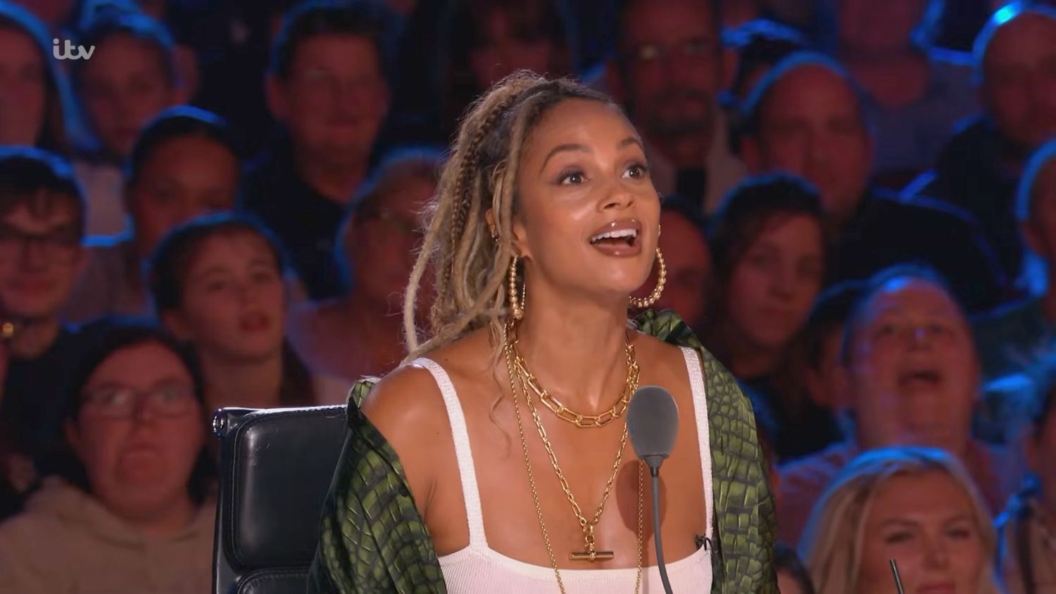 Alesha Dixon smiling as a judge on Britain's Got Talent, wearing a white top and green cardigan with gold jewelry.