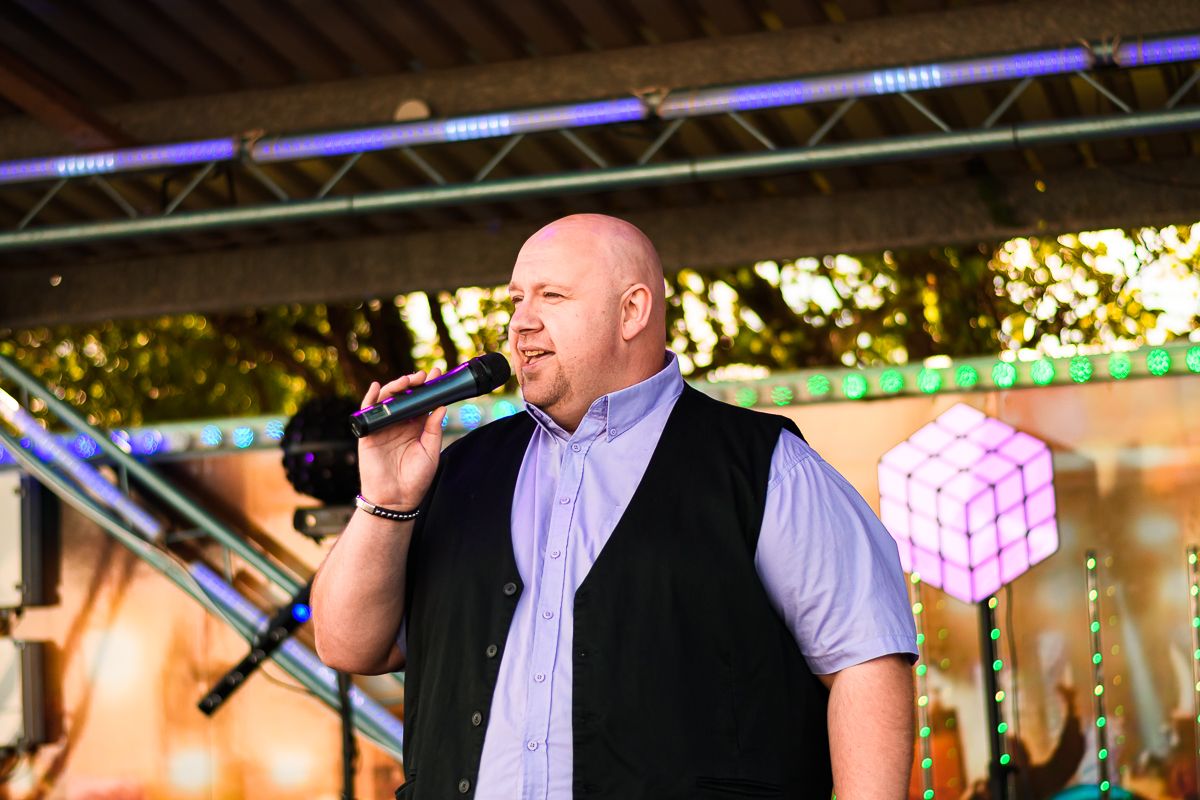 Bald male singer performing on outdoor stage at night, holding microphone, wearing black vest over blue shirt.