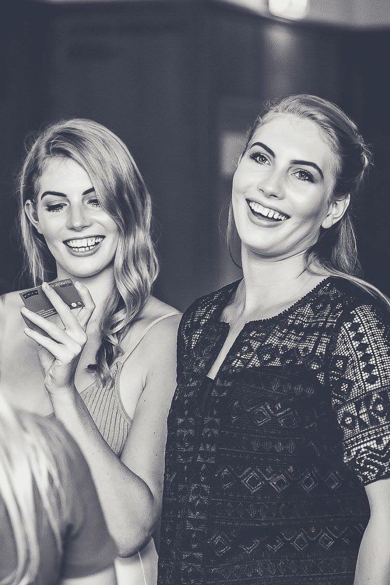 Two smiling blonde women pose together at an evening event, one holding a smartphone.