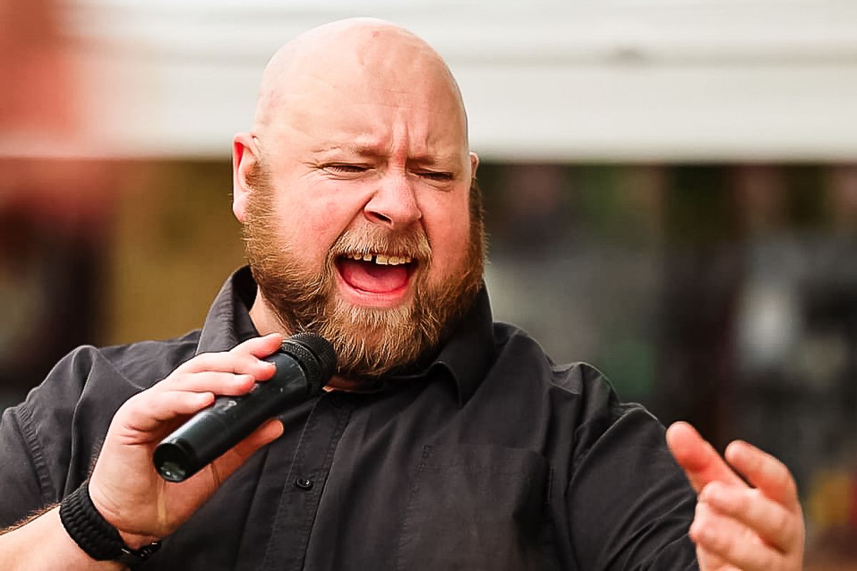 Bald bearded man singing passionately into a microphone outdoors, wearing a black shirt.