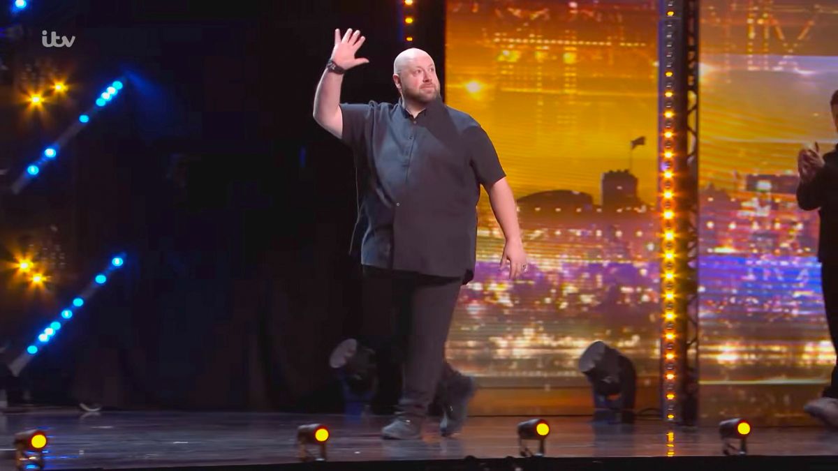 Overweight man waves to audience on Britain's Got Talent stage under bright spotlights.