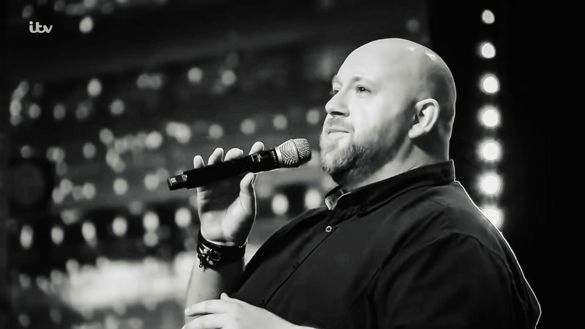 Bald bearded man singing into microphone on stage in black and white performance photo.