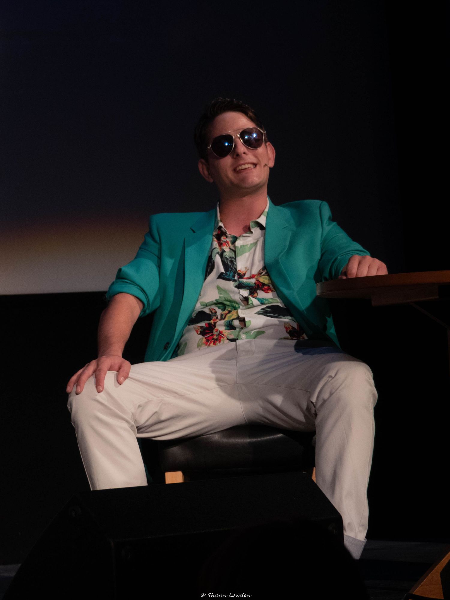 Performer in teal blazer and white pants lounging on stage during theatrical production.