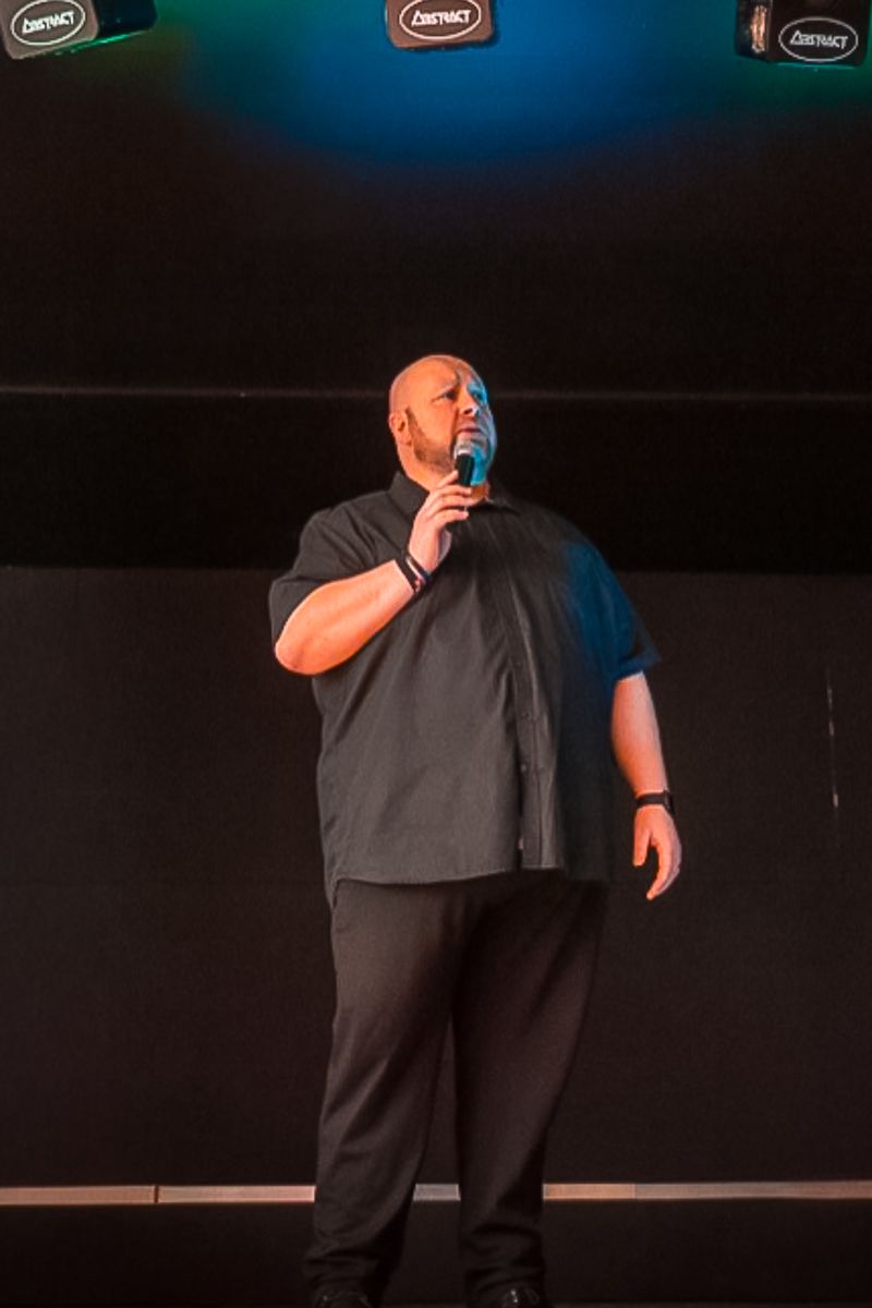 Bald man in black outfit performing stand-up comedy on stage, holding a microphone under colorful stage lighting.