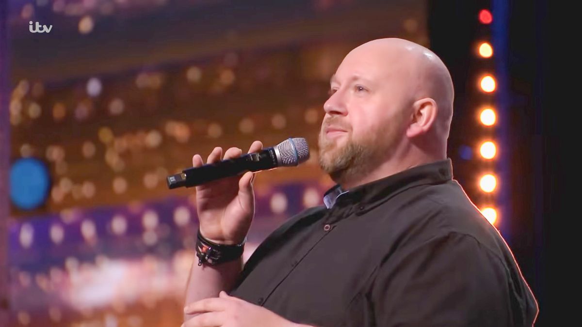 Bald man singing into a microphone on the Britain's Got Talent ITV stage under bright lights.