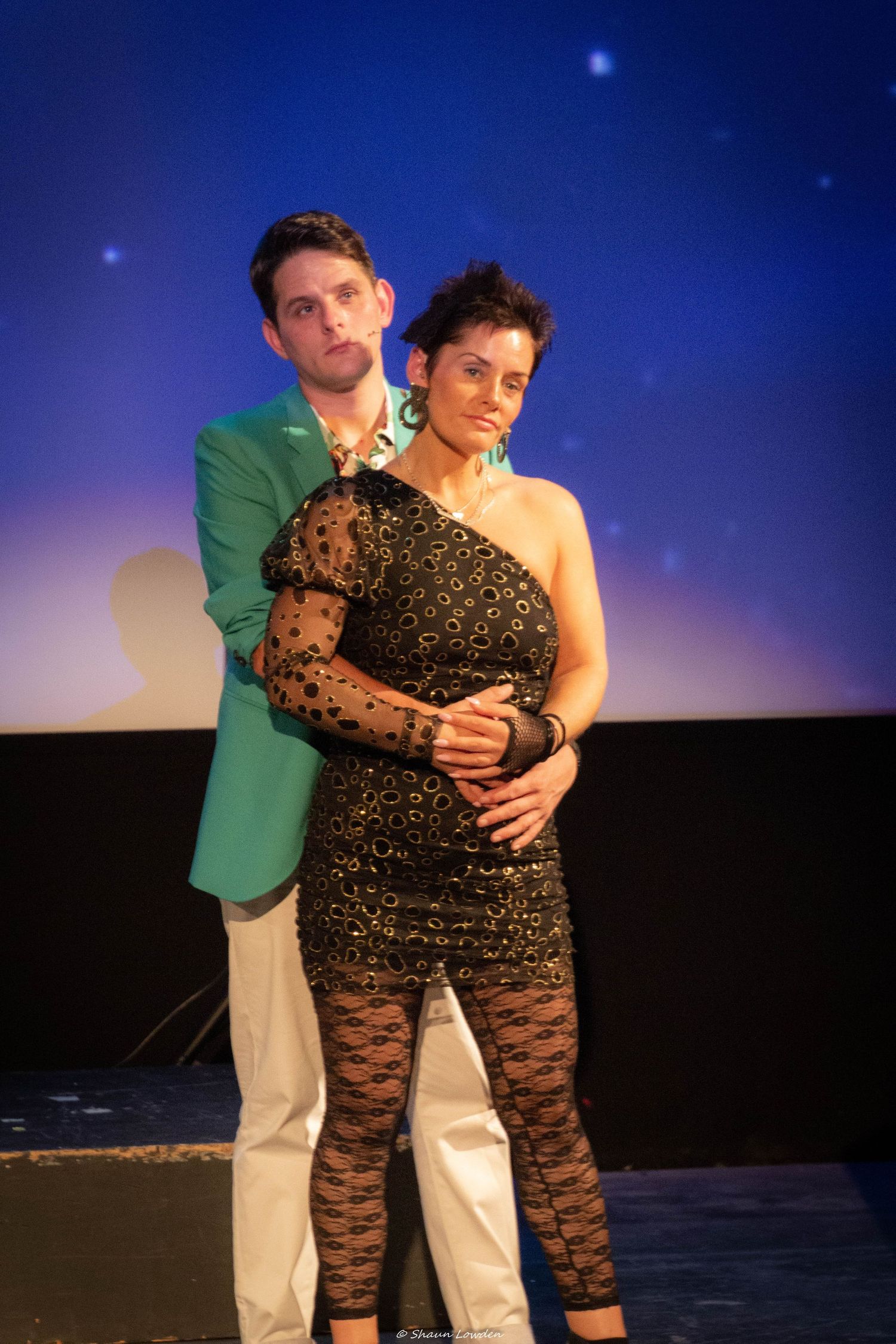 Performers in a theatrical embrace against a dark blue backdrop during a stage performance.