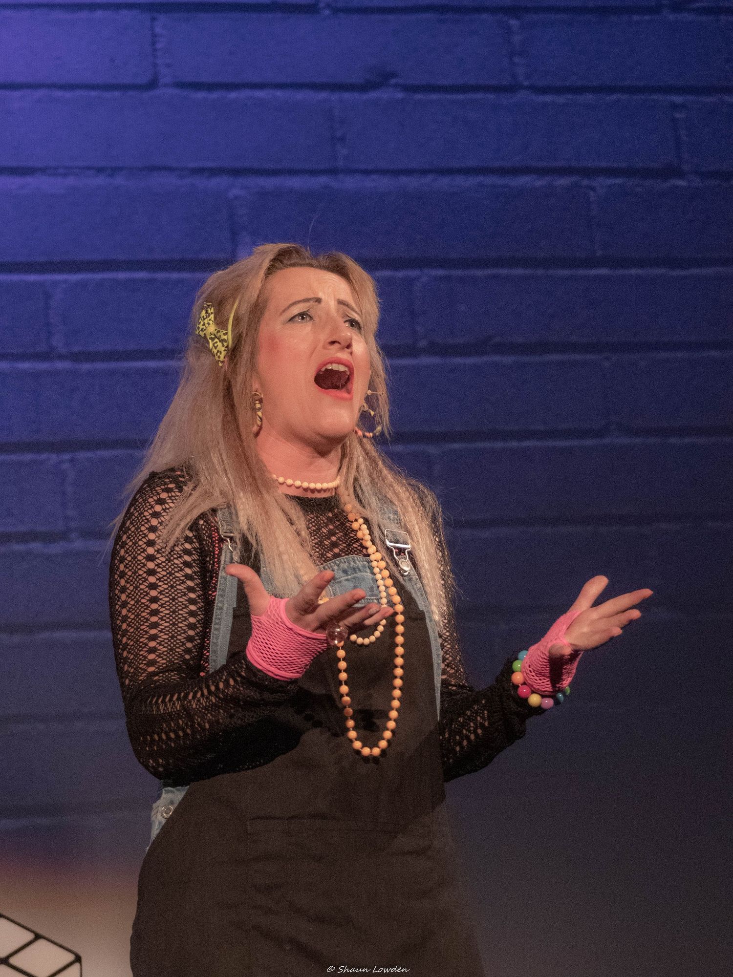 A performer in black mesh clothing gestures dramatically against a blue-lit brick wall.