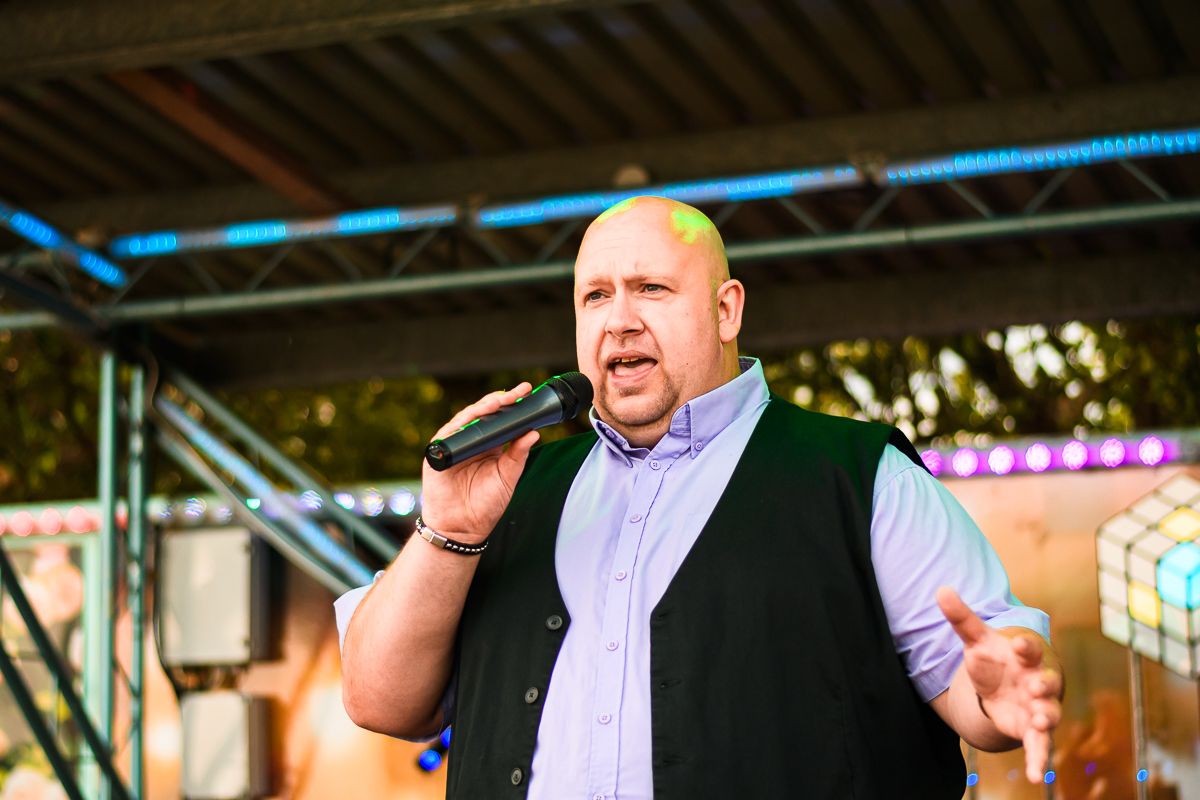 Bald man singing into microphone on outdoor stage, wearing black vest over light blue shirt at evening event.