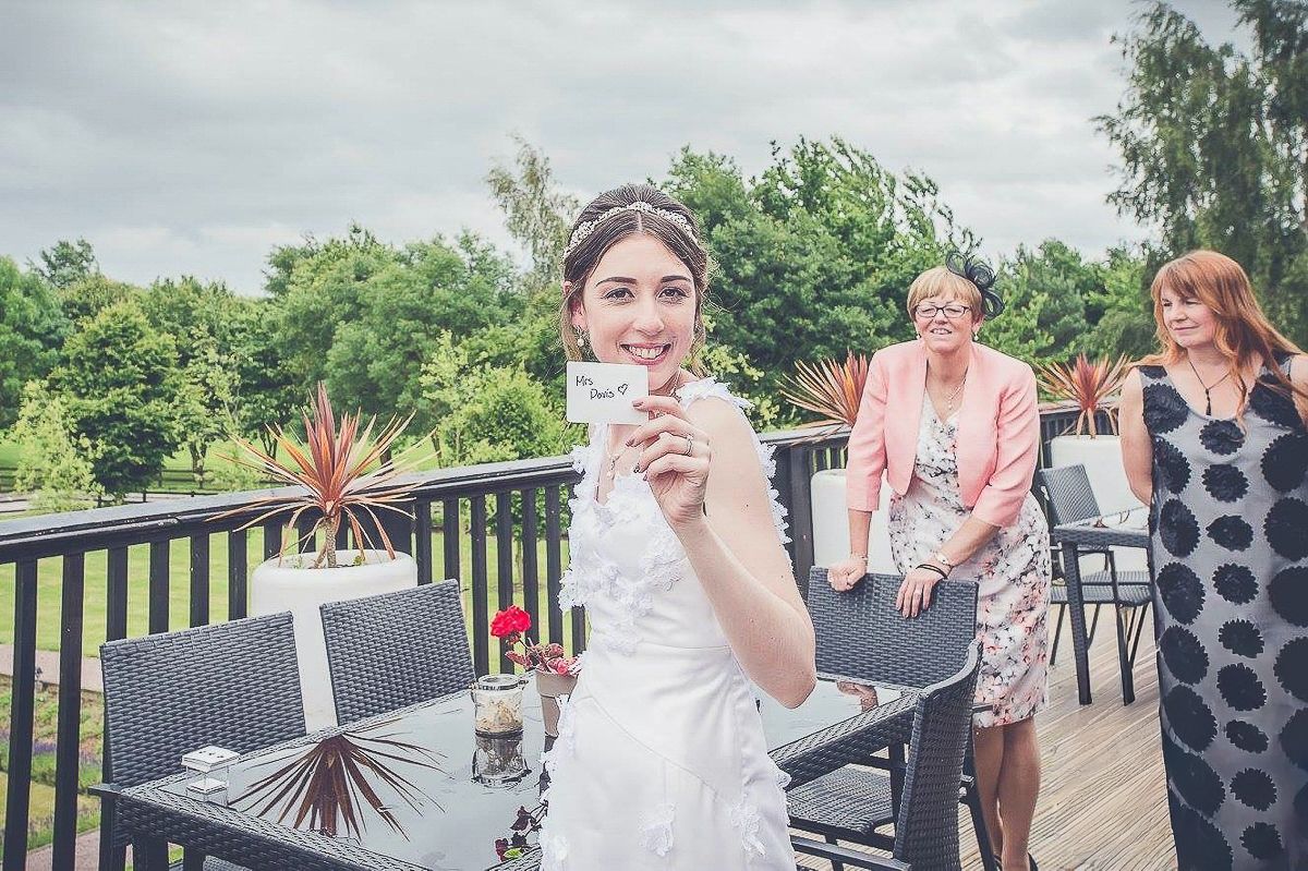 A bride holding a card outdoors on a deck with wedding guests smiling in background.