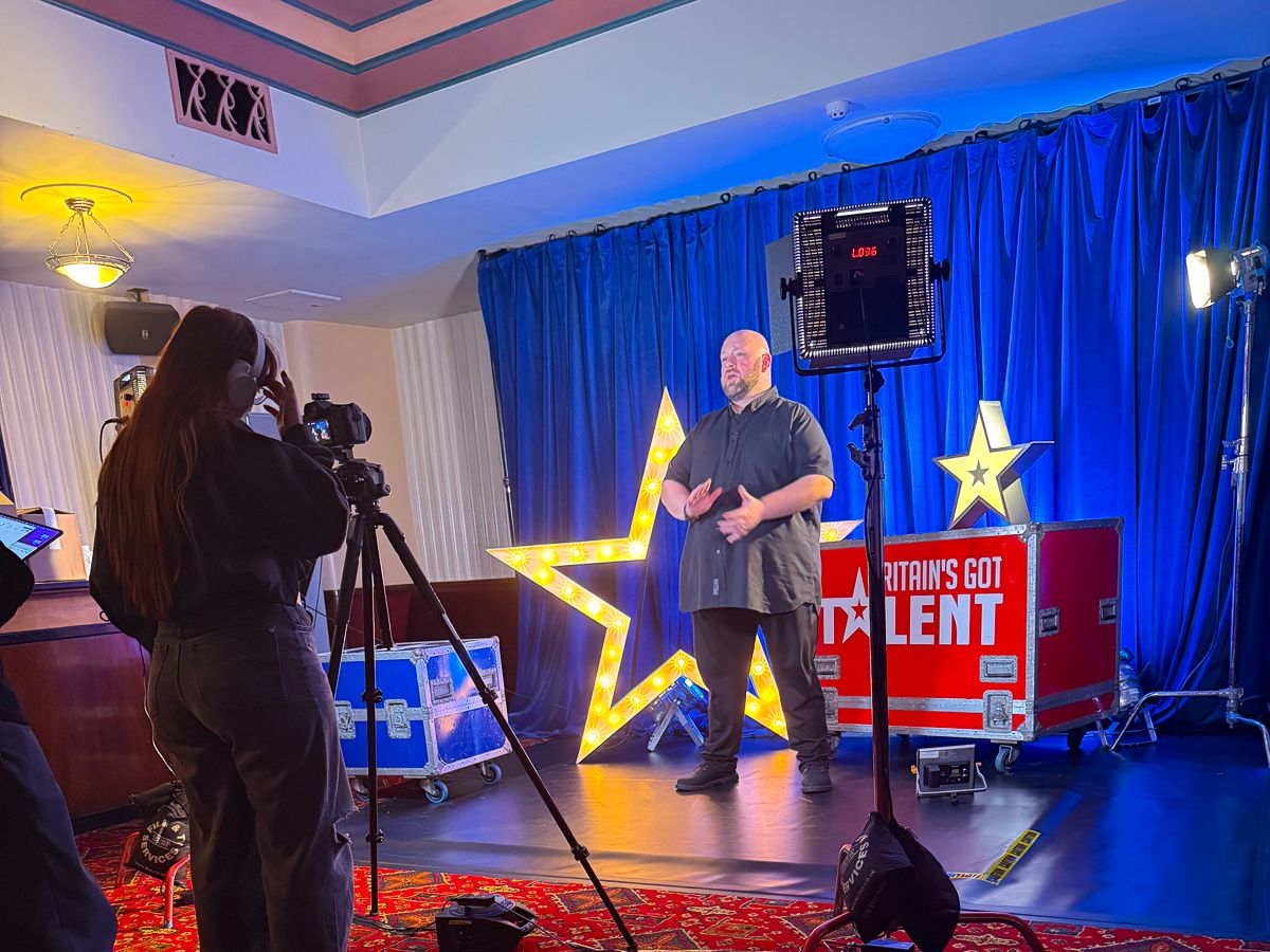 Man performs on Britain's Got Talent stage set with gold stars, blue curtains, and a camera operator filming.
