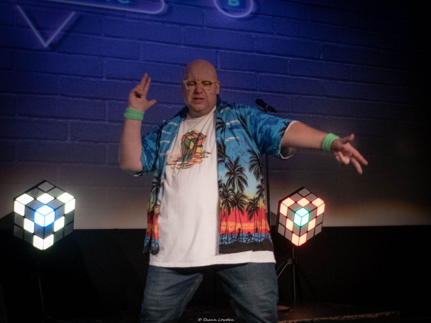 Performer in tropical print shirt gesturing dramatically on stage with blue lighting.