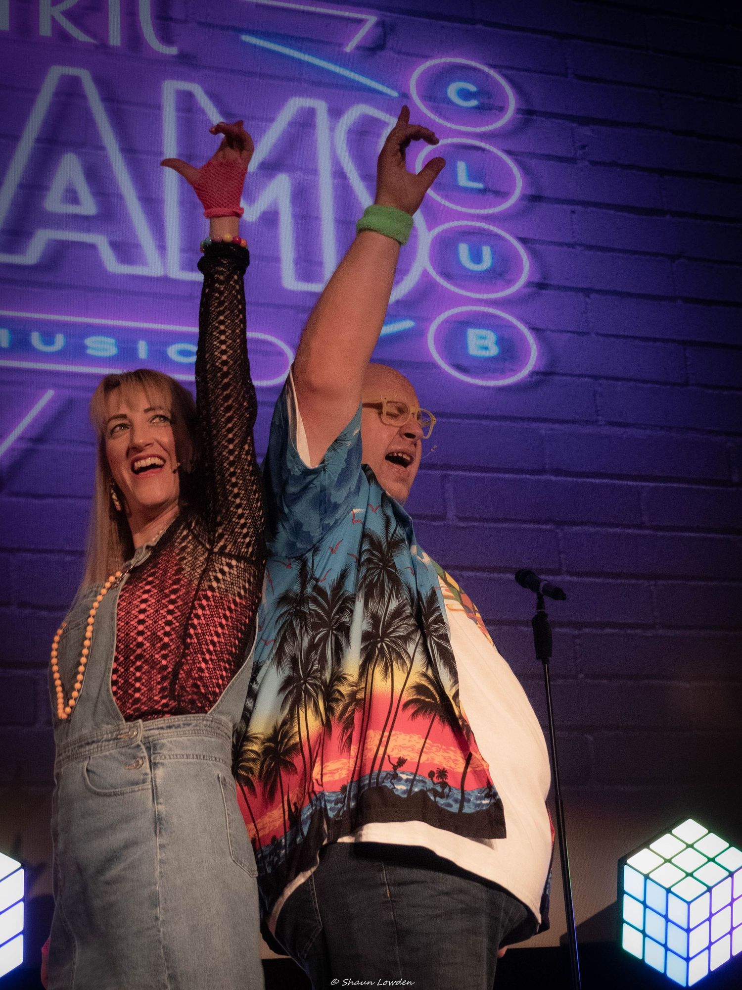 Two performers raising arms triumphantly on stage with neon lighting.
