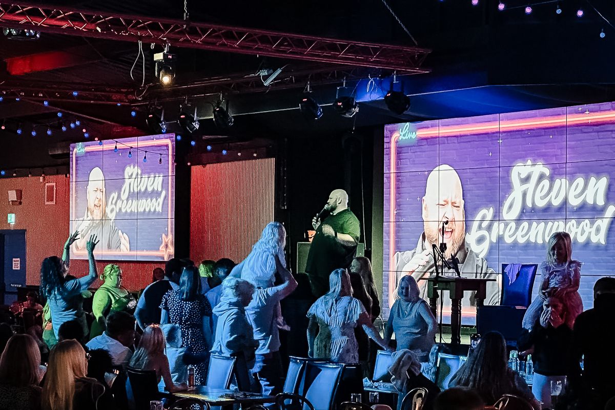 Concert crowd stands before large video screens displaying branding while stage lights glow in red and blue tones.