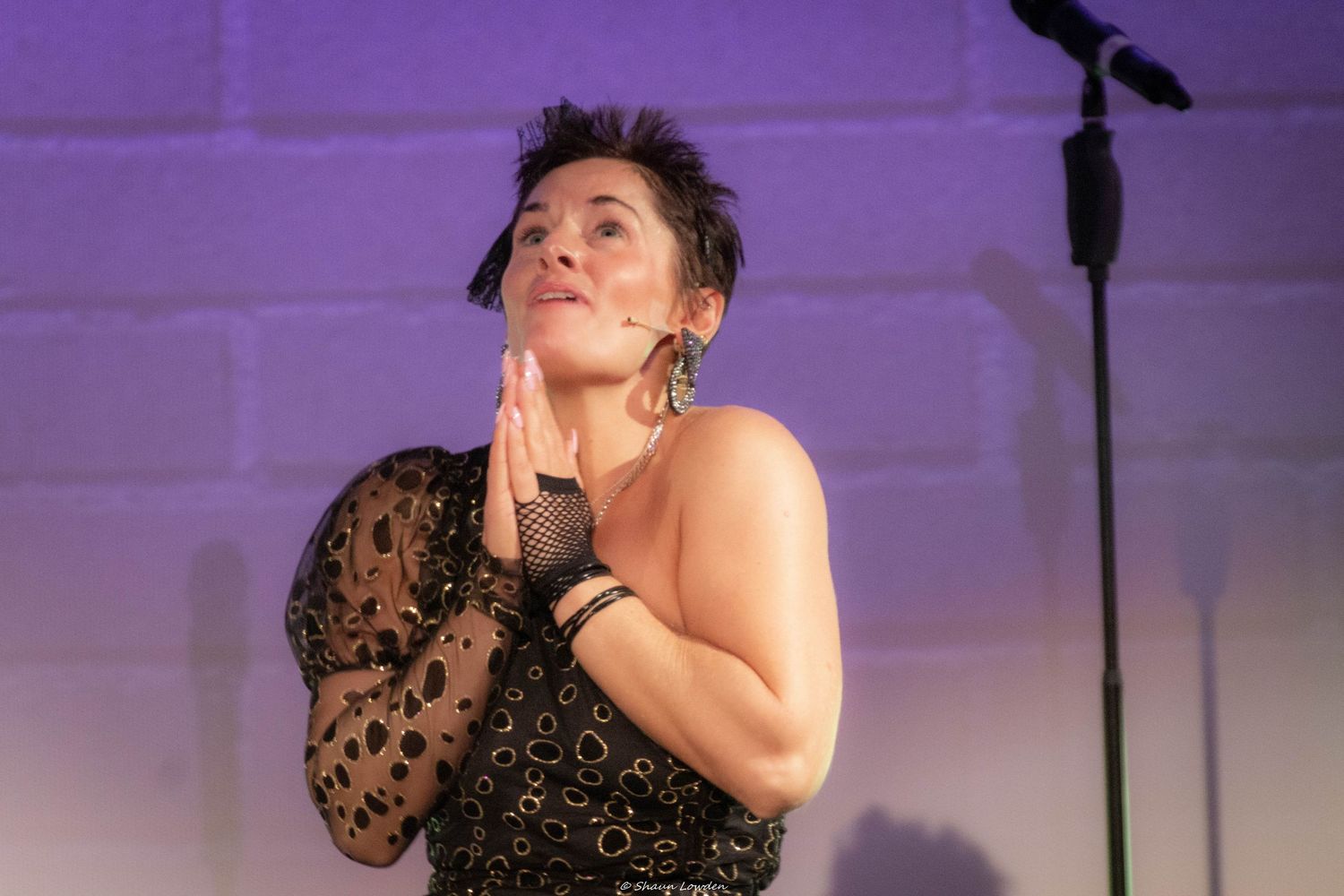 Performer in black lace dress striking dramatic pose on purple-lit stage.