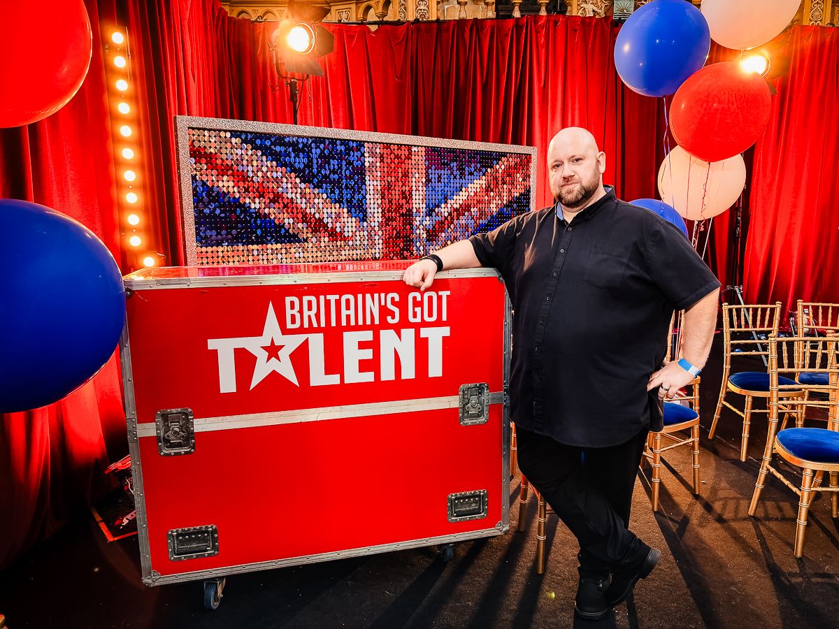 Man posing next to a red Britain's Got Talent branded flight case on stage with red curtains and balloons.