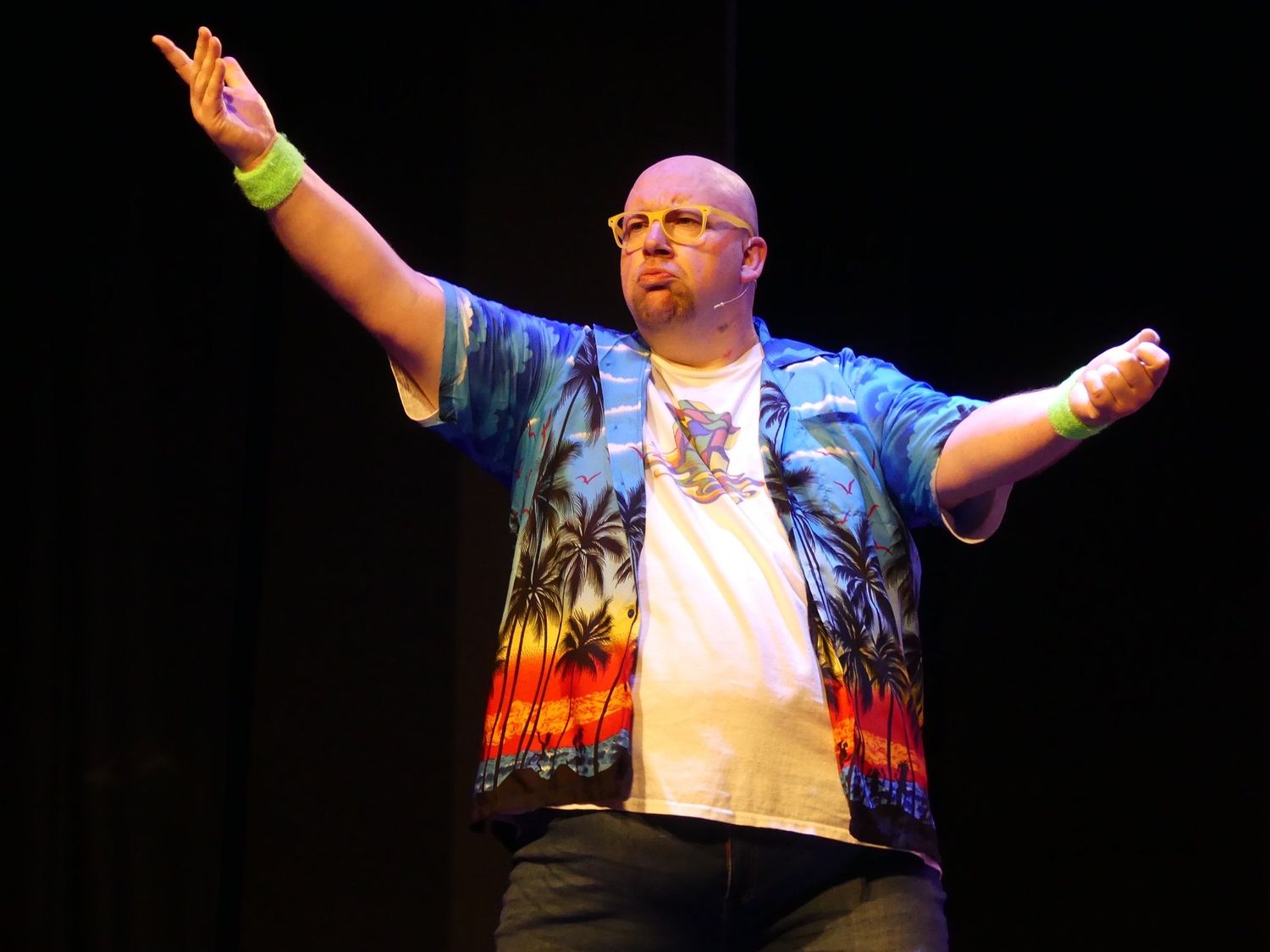Performer in patterned jacket with arms raised on dark stage with dramatic lighting.