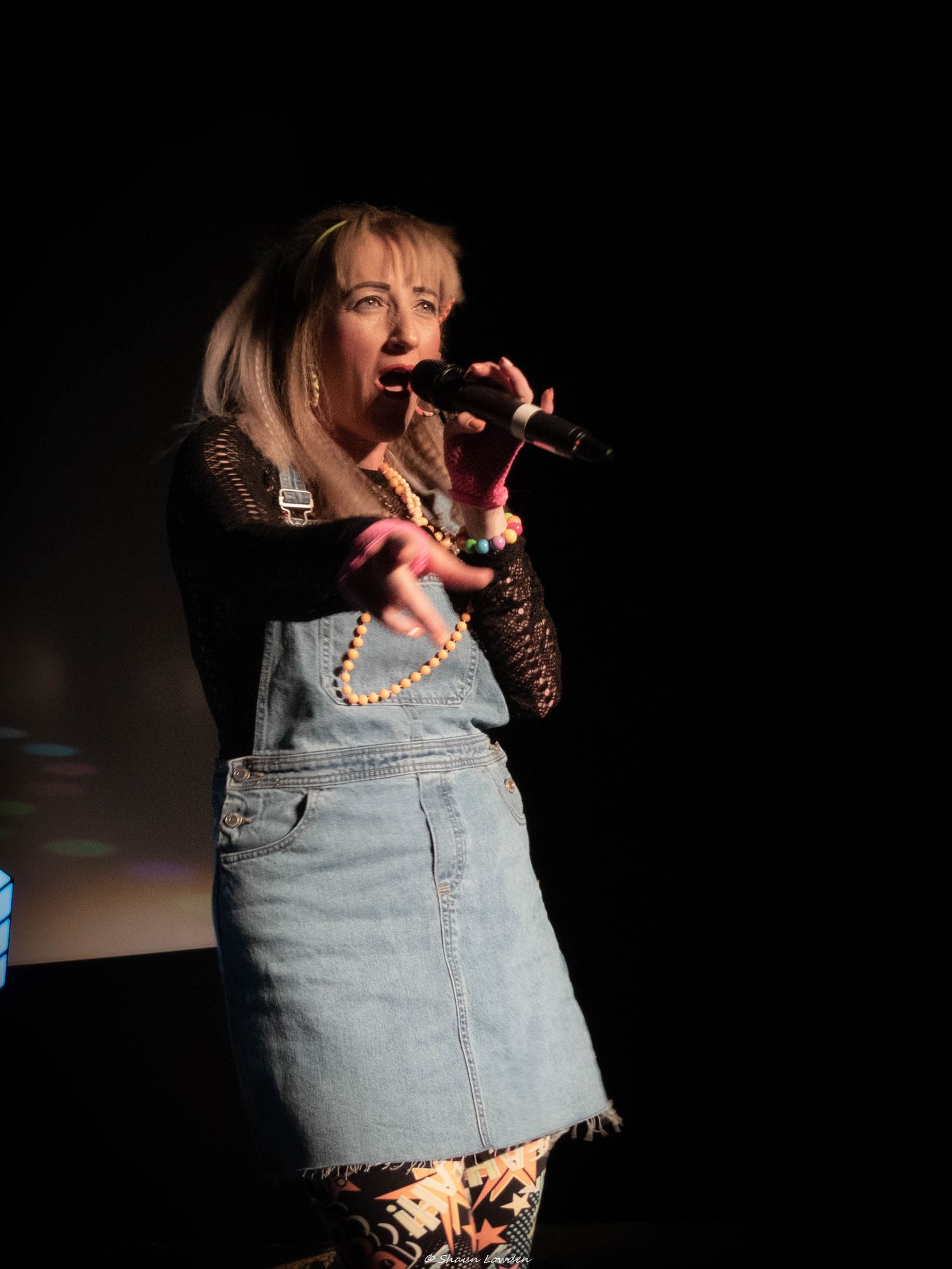 Singer in denim overalls dress performing on dark stage with microphone.