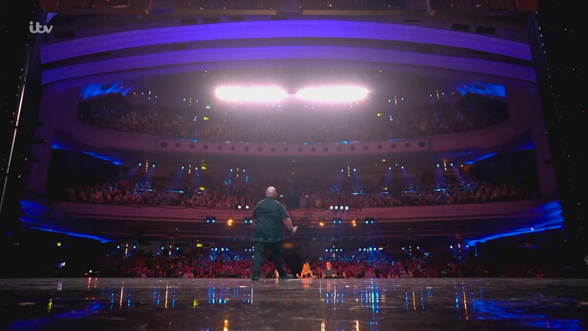 Performer stands on stage at a grand theater with tiered seating, colorful stage lighting, and a large audience.