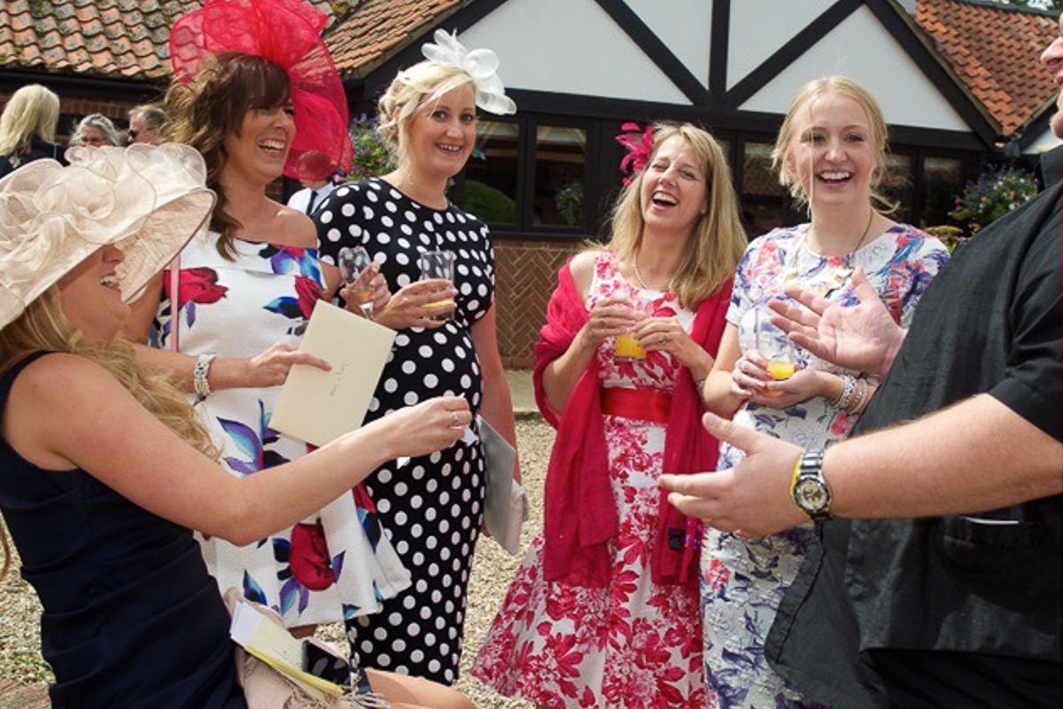 Elegantly dressed women in colorful floral dresses and fascinators laughing together at a garden party event.