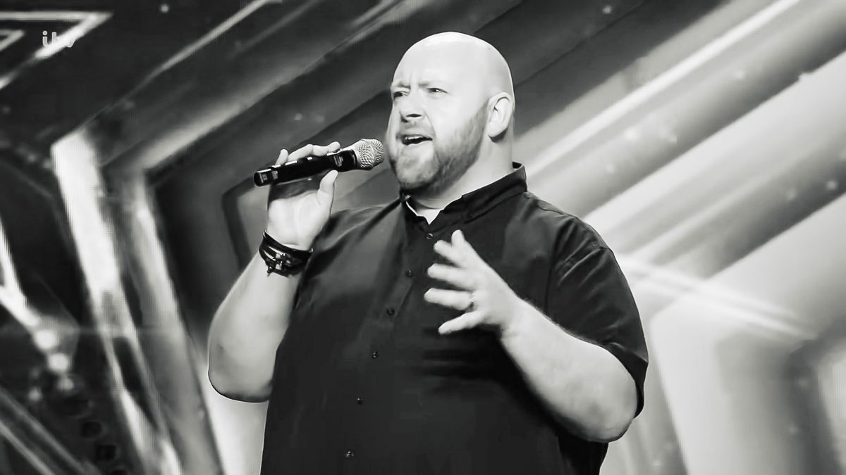 Man in black shirt singing passionately into a microphone on a dramatic stage with X-shaped lighting.