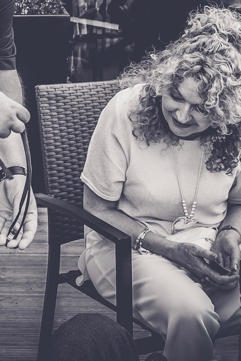 Black and white photo of a smiling woman with curly hair sitting in a wicker chair, wearing a necklace and bracelet.
