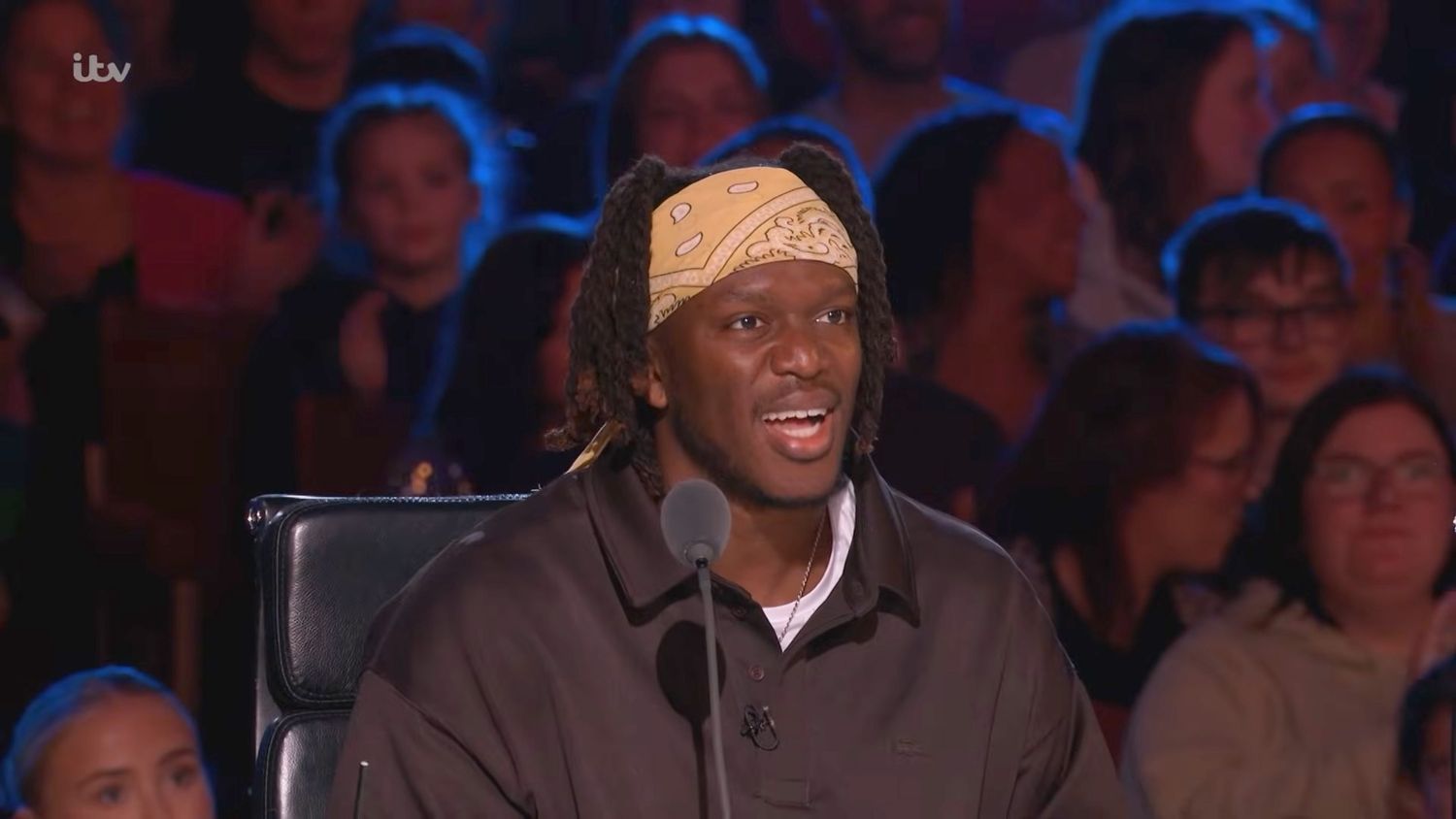 A man wearing a yellow bandana and dark jacket sits as a judge on a TV talent show with audience behind him.