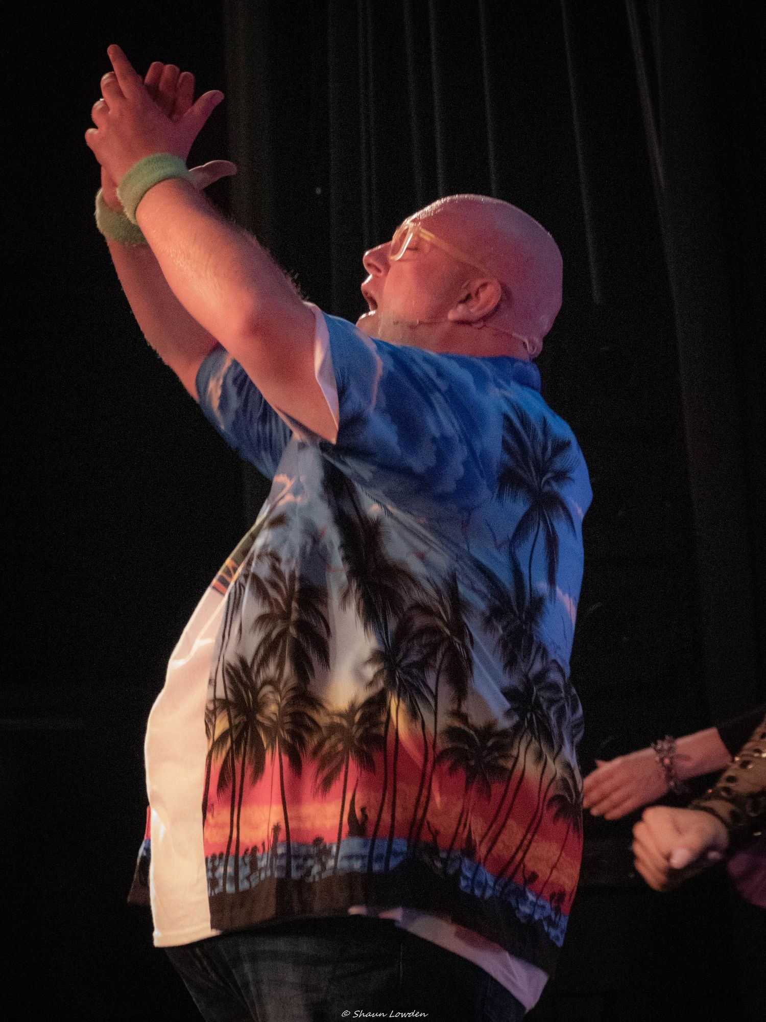 Performer in a tropical print shirt raises their arms during an energetic stage performance with colorful lighting.