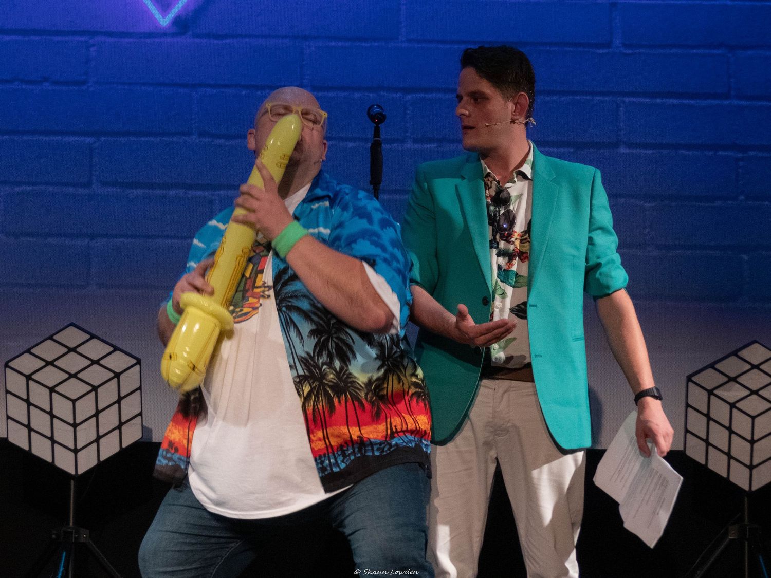 Performers in casual attire interact during a stage show with geometric backdrop elements.