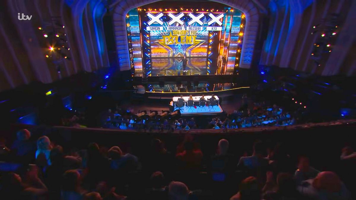 A packed theater audience watches a colorful stage performance with bright lights and signage at a live TV show taping.