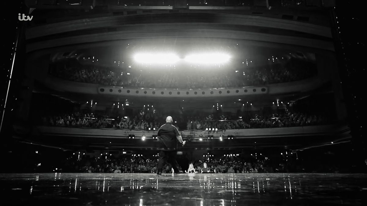 Black and white photo of a performer on stage facing a packed theater audience under a bright spotlight.
