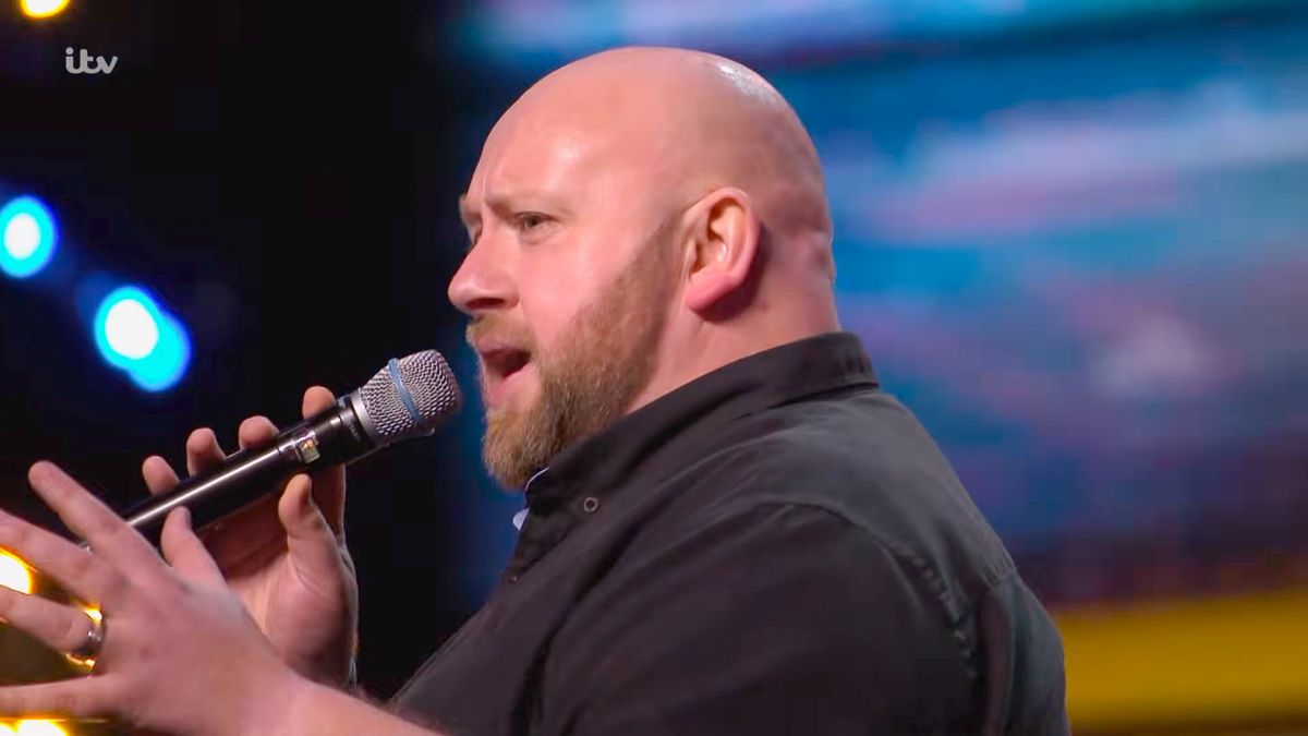 Bald man singing passionately into a microphone on a talent show stage with dramatic lighting.