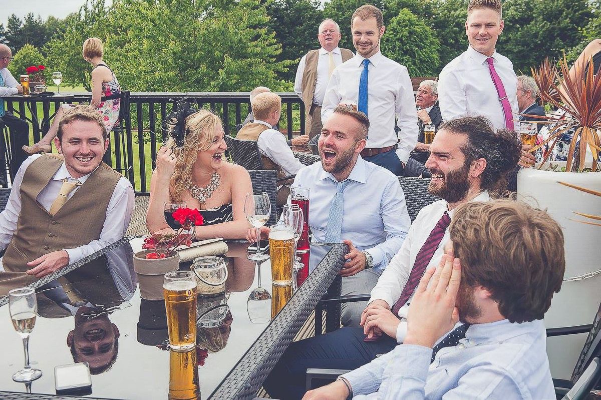 Guests laughing and socializing at an outdoor wedding reception, seated at a glass table with drinks.