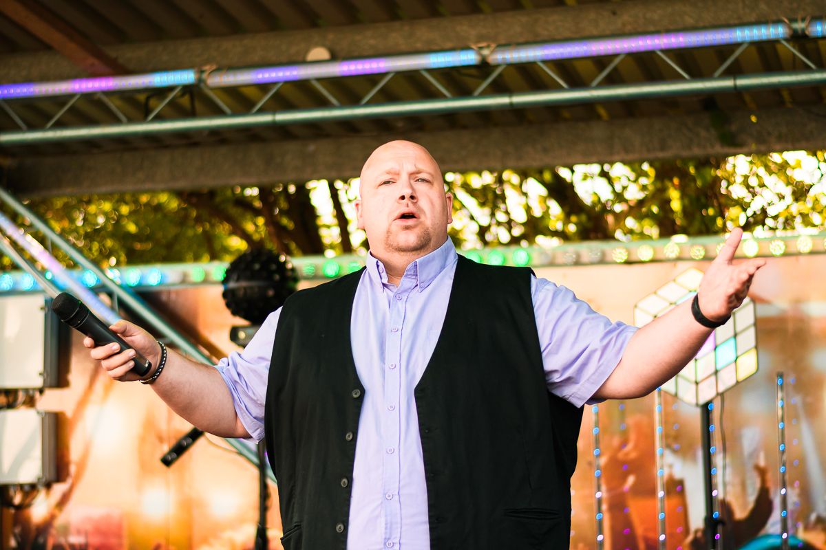 Bald man in black vest performing on outdoor stage with arms outstretched, singing at a festival event.