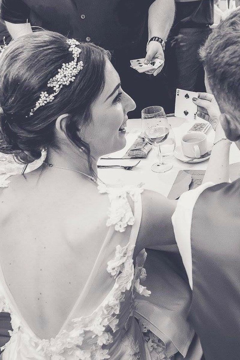 A bride in a lace wedding dress watching a card trick performed at her reception.