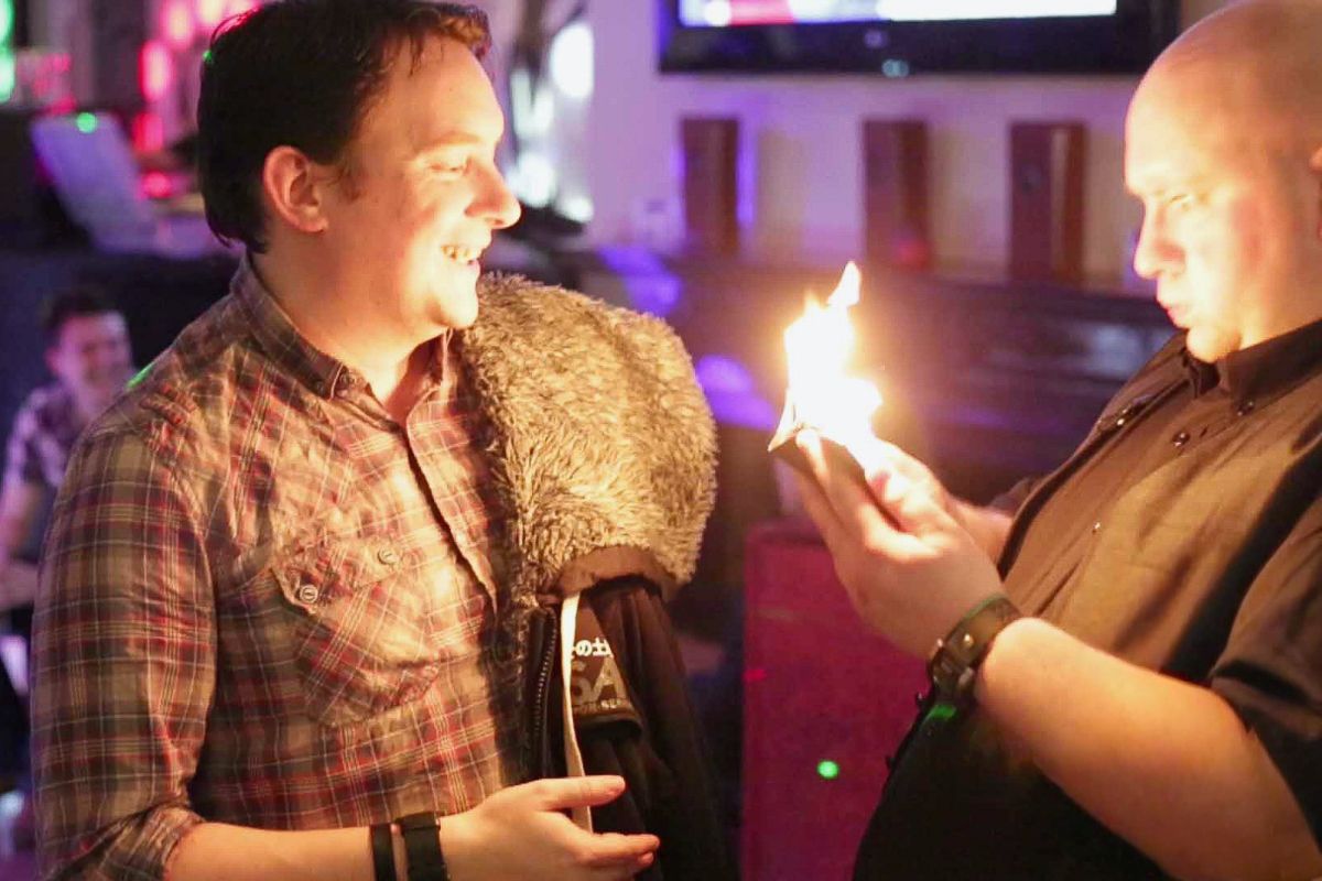 Two men at a bar, one performing a fire magic trick with a hat.