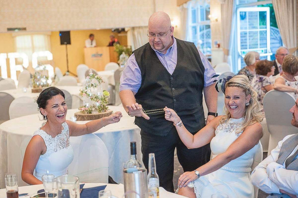 A magician entertains two women in white dresses at an elegant wedding reception venue.
