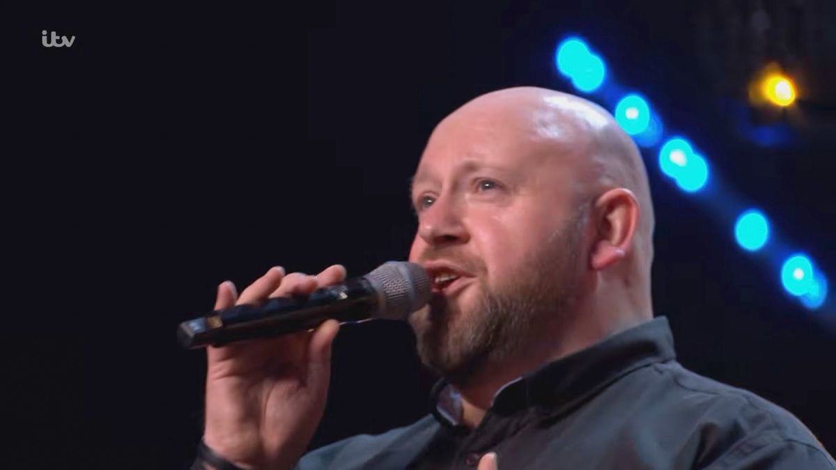 Bald man with beard singing into microphone on ITV stage with blue lights in background.