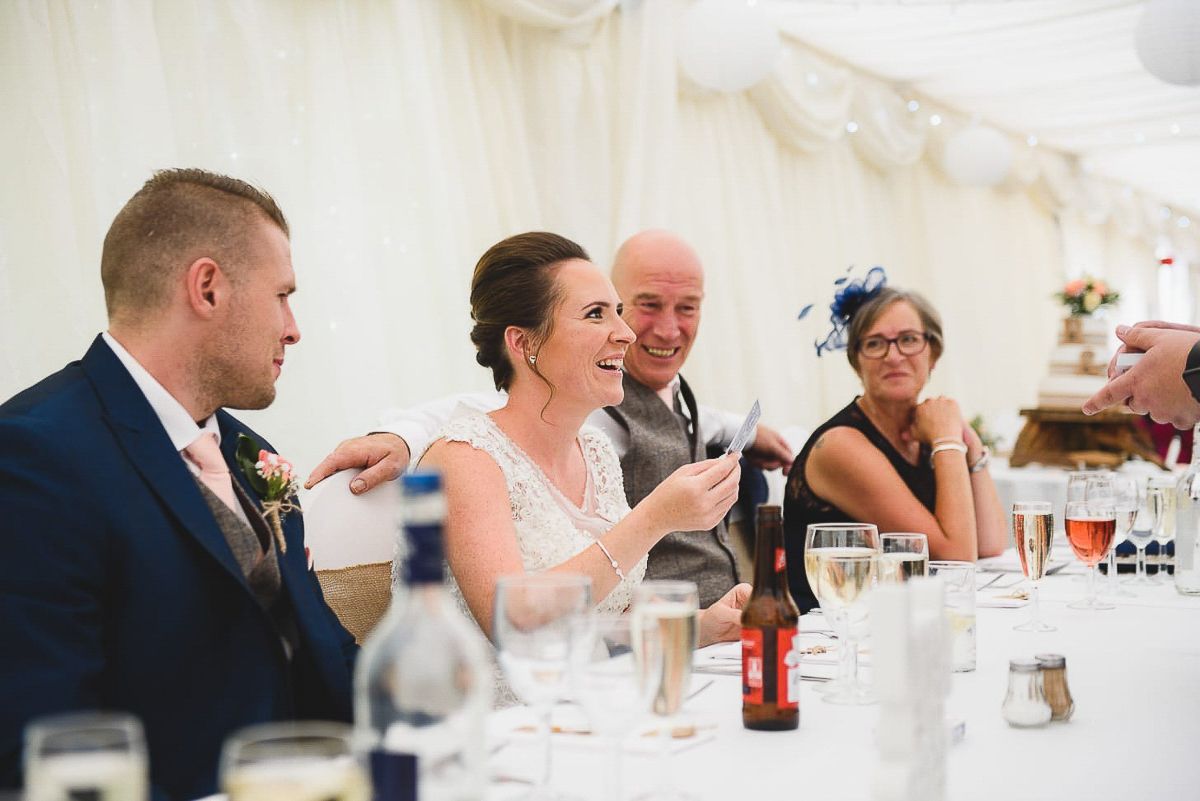 Bride and groom laughing with guests at elegant wedding reception dinner table in white marquee.